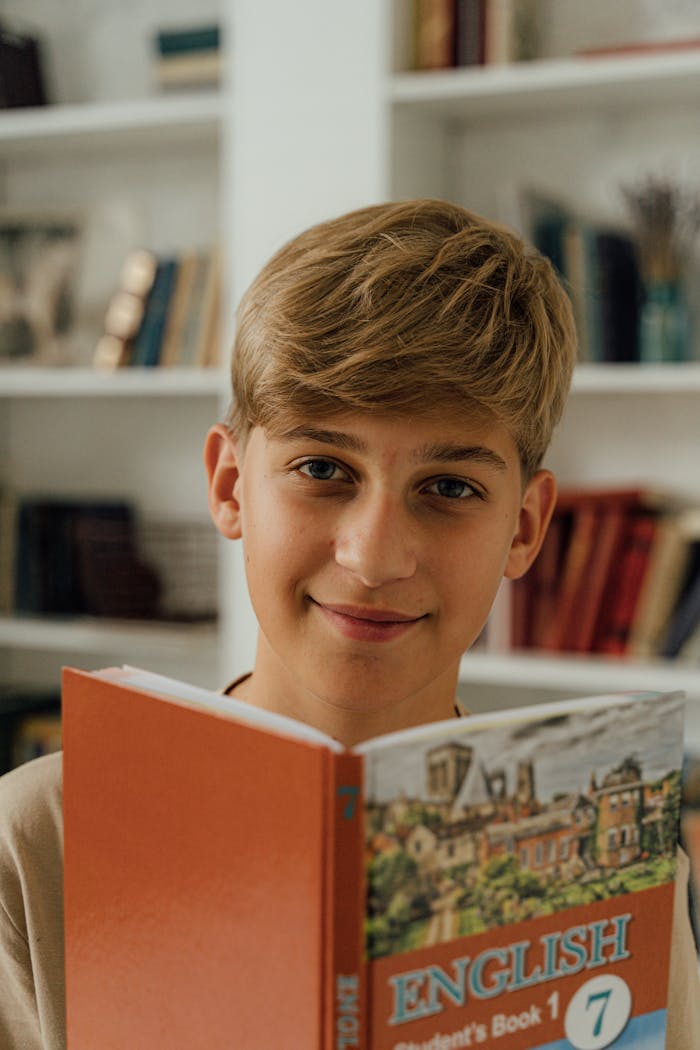 about-04 Young boy smiling while reading an English textbook indoors in a library setting.