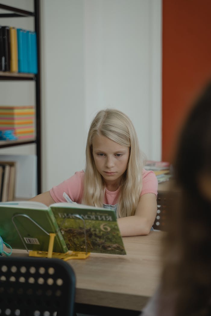 services-01 Blonde girl intently reads a book at school desk, embodying education and learning.